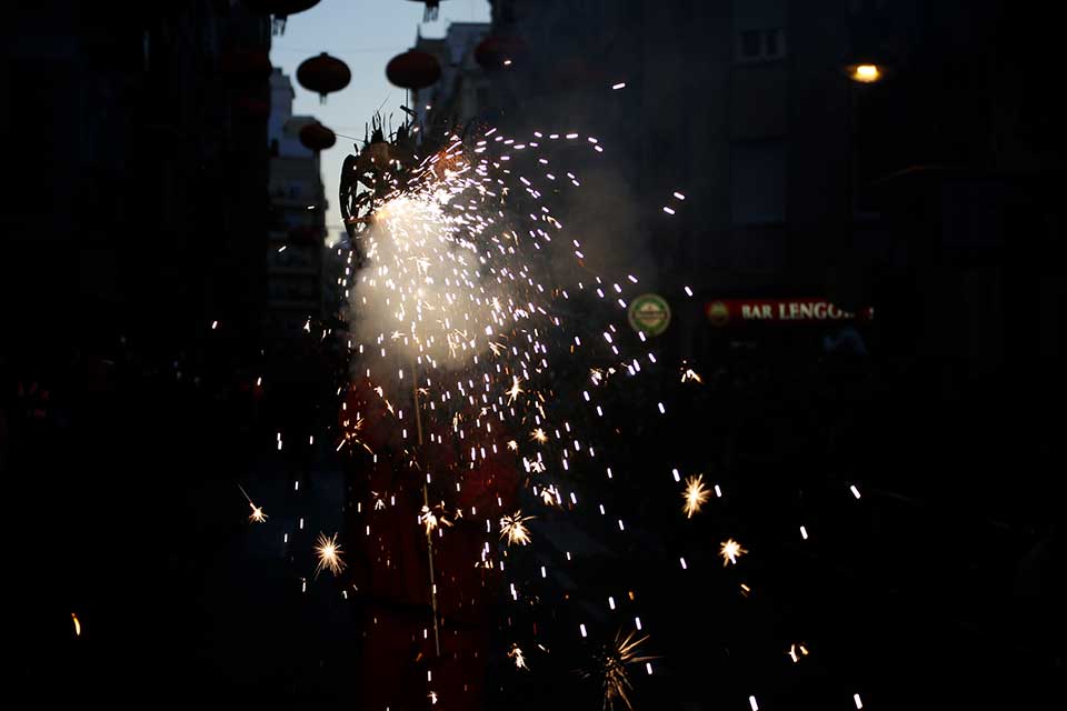 A photograph of a firework erupting at dusk. The darkening sky is just visible in the gap between two buildings.